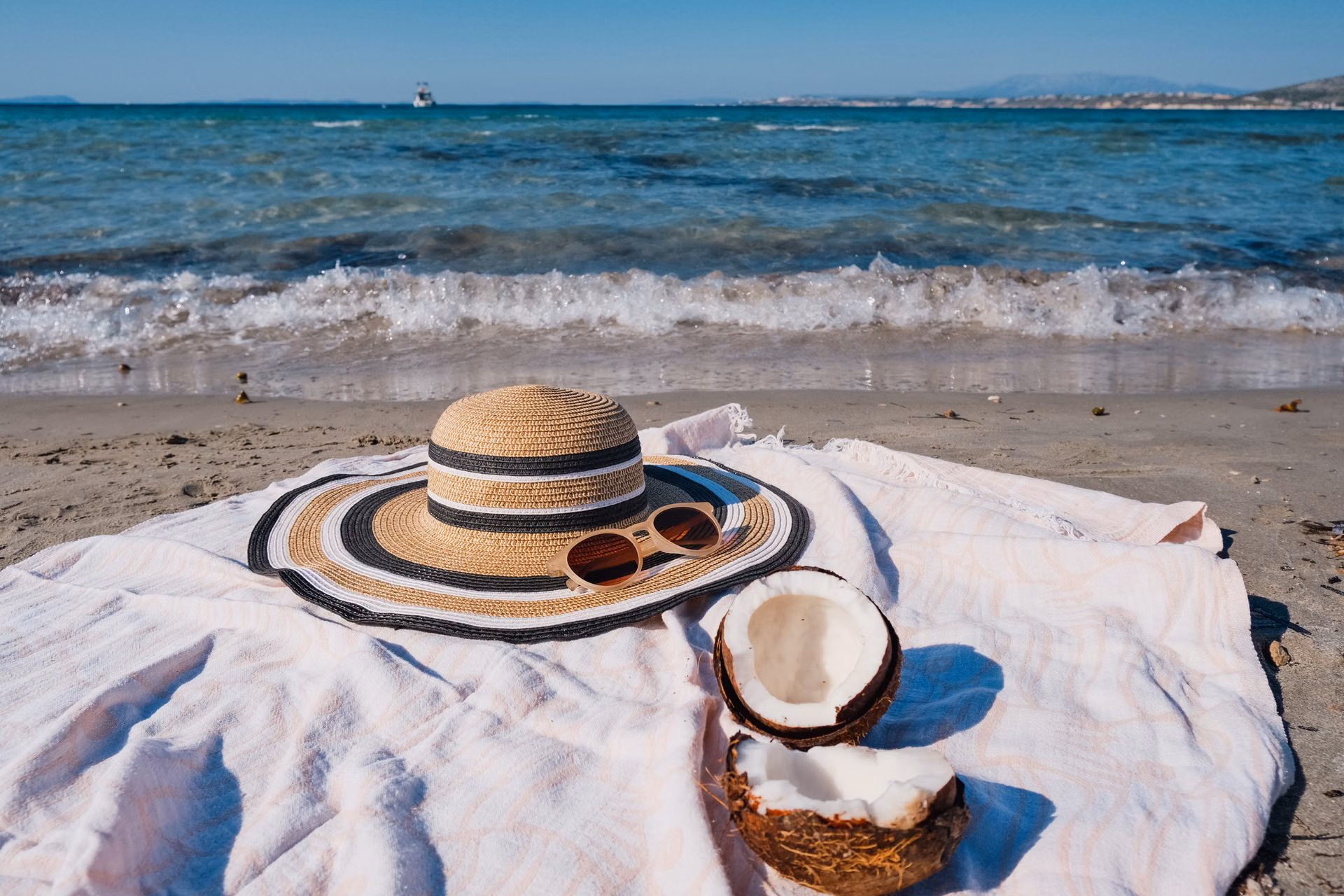A straw sun hat, sunglasses and a coconut on a towel by the sea
